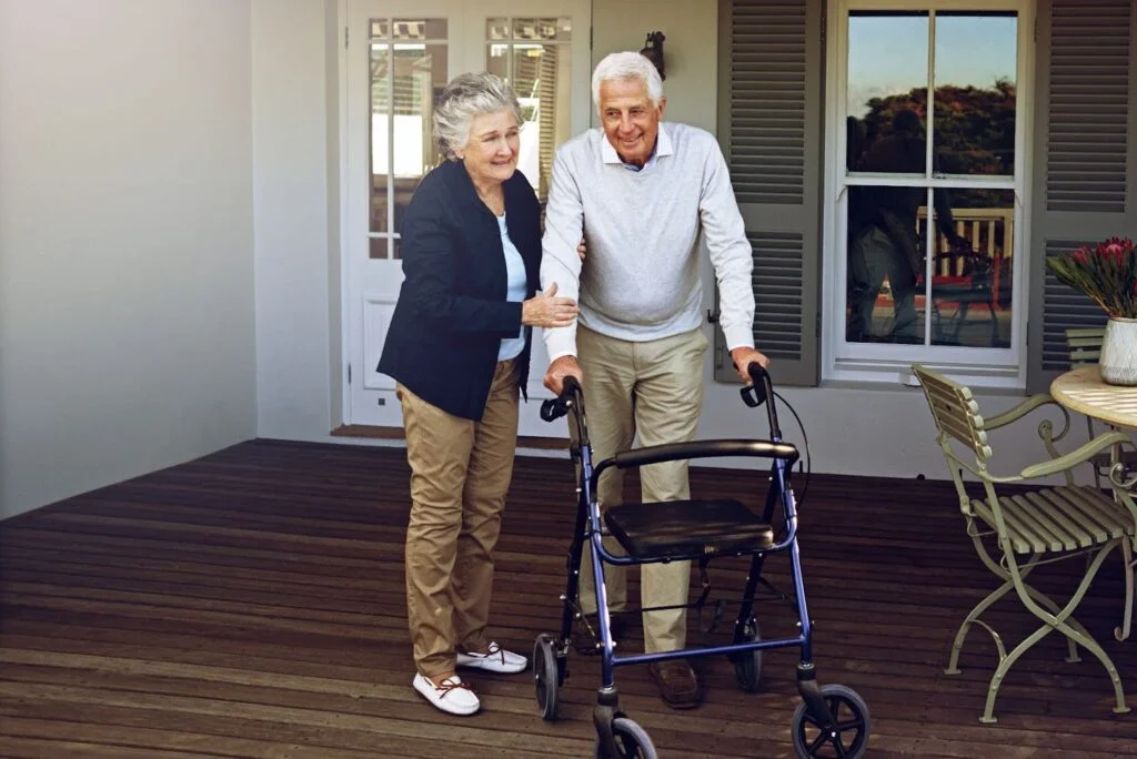 Older man using a large wheel walker on a porch with caregiver support